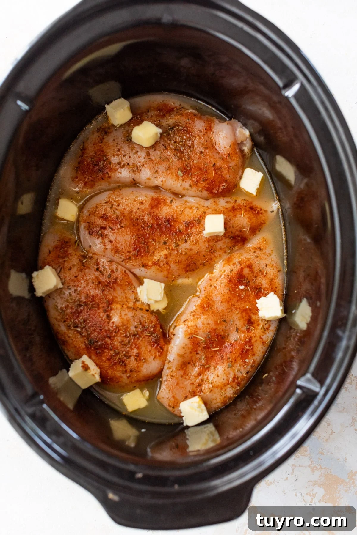 Tender Slow-Cooker Chicken Breast Recipe for Quick Meals 5 raw seasoned chicken breasts in a slow cooker with broth and pats of butter.