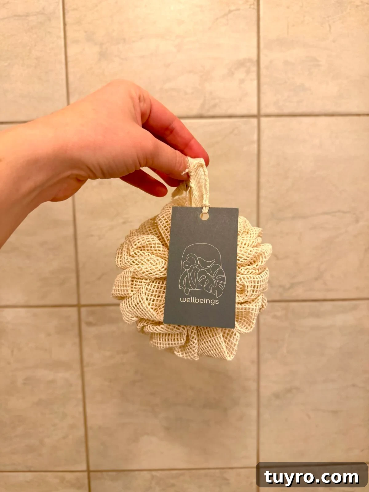 Hand holding a natural mesh loofah with a “wellbeings” tag against a tiled bathroom wall.