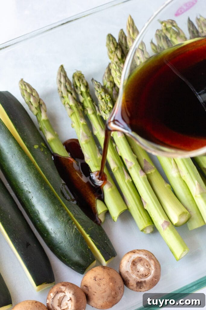 A measuring cup pouring a balsamic marinade over raw asparagus and zucchini in a glass dish.
