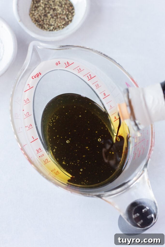 Pouring balsamic vinegar into a measuring cup with oil in it that's on top of a white background next to small bowls of salt and black pepper.
