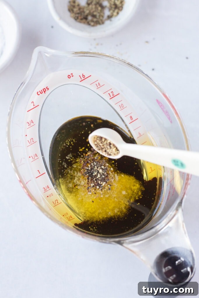 A small measuring spoon dumping ground black pepper into a measuring cup that has oil, vinegar, and salt already in it.