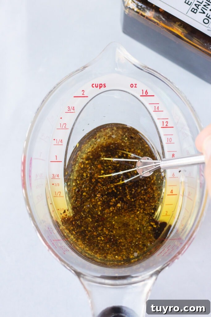 A hand using a small whisk to stir together vinegar, oil, salt, and pepper in a plastic measuring cup that's sitting on a white surface.