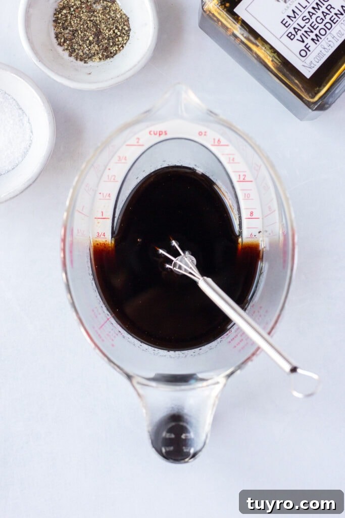 Top down shot of a plastic measuring cup with a balsamic marinade in it, with a whisk resting in it as well. Toward the top of the frame are two small white bowls with salt and pepper, and a bottle of balsamic vinegar, all on a white background.