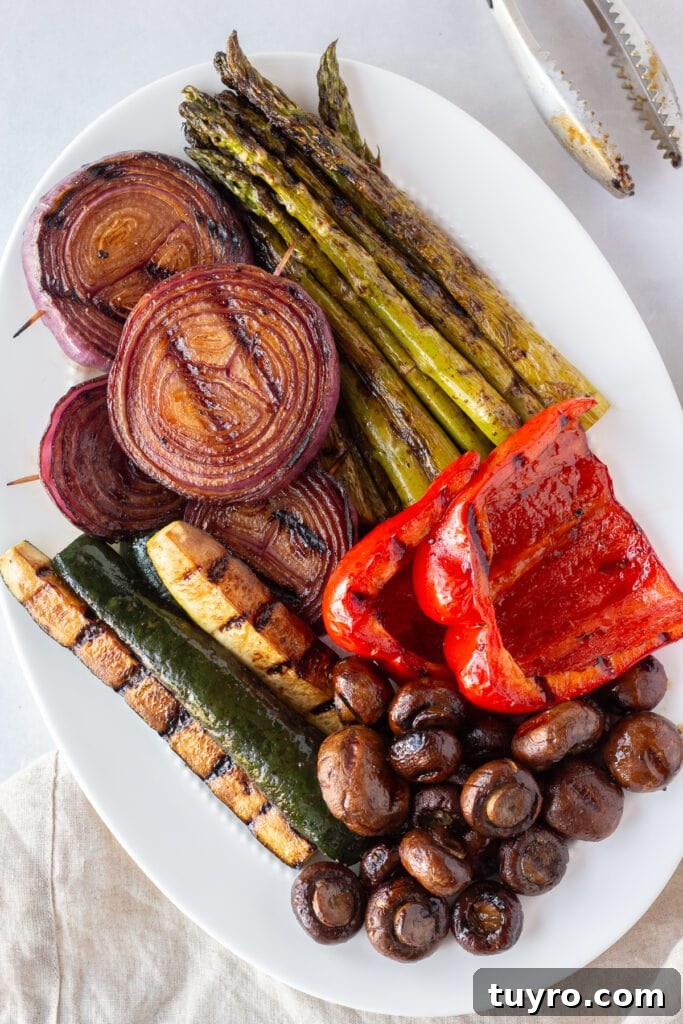 Top down shot of marinated and grilled red onion, zucchini, asparagus, mushrooms, and bell pepper on a white oval platter next to a pair of tongs and a tan napkin, all on a white background.