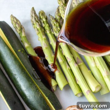 A measuring cup pouring a balsamic marinade over raw asparagus and zucchini in a glass dish.