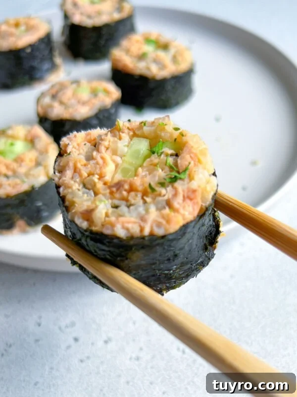 A pair of chopsticks holds a sushi roll with seaweed, rice, and a filling of salmon and cucumber. Several similar sushi pieces are arranged on a white plate in the background.