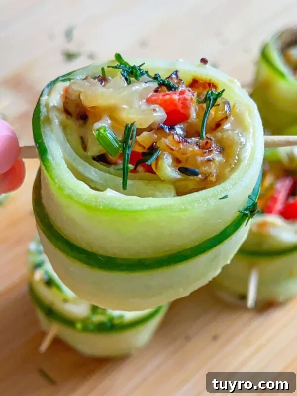 A hand holds a skewer with a zucchini roll-up stuffed with melted cheese, red peppers, and herbs. Other zucchini roll-ups share the wooden surface, offering fresh inspiration for snacks like Cucumber Salmon Boats.