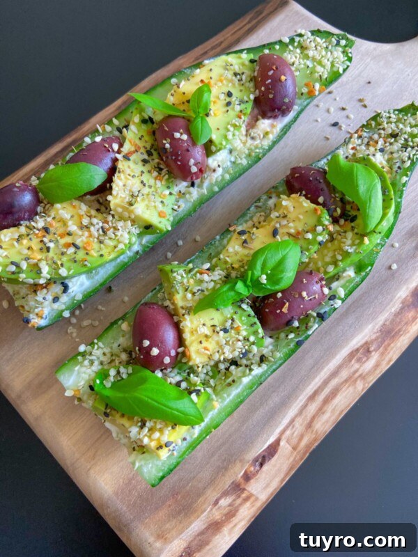 Two Egg Salad Cucumber Boats topped with sliced avocado, Kalamata olives, fresh basil leaves, hemp seeds, and seasoning, served on a wooden board against a dark background.