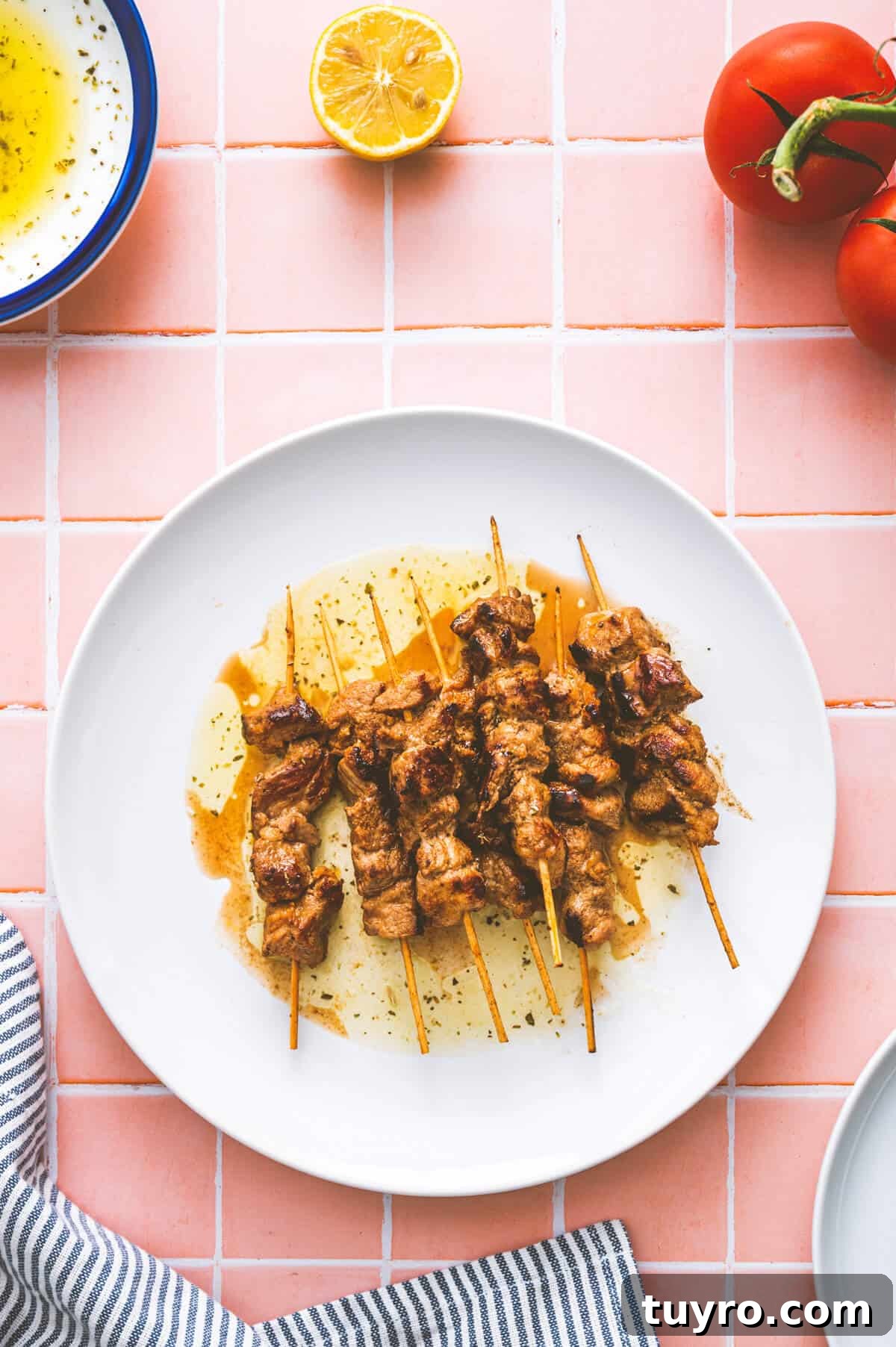 Souvlaki Explained: Your Guide to Greece’s Iconic Street Food 4 A white plate with grilled meat skewers drizzled with oil sits on a pink tiled surface. Nearby are a halved lemon, fresh tomatoes on the vine, a bowl of oil, and a striped cloth serviette.