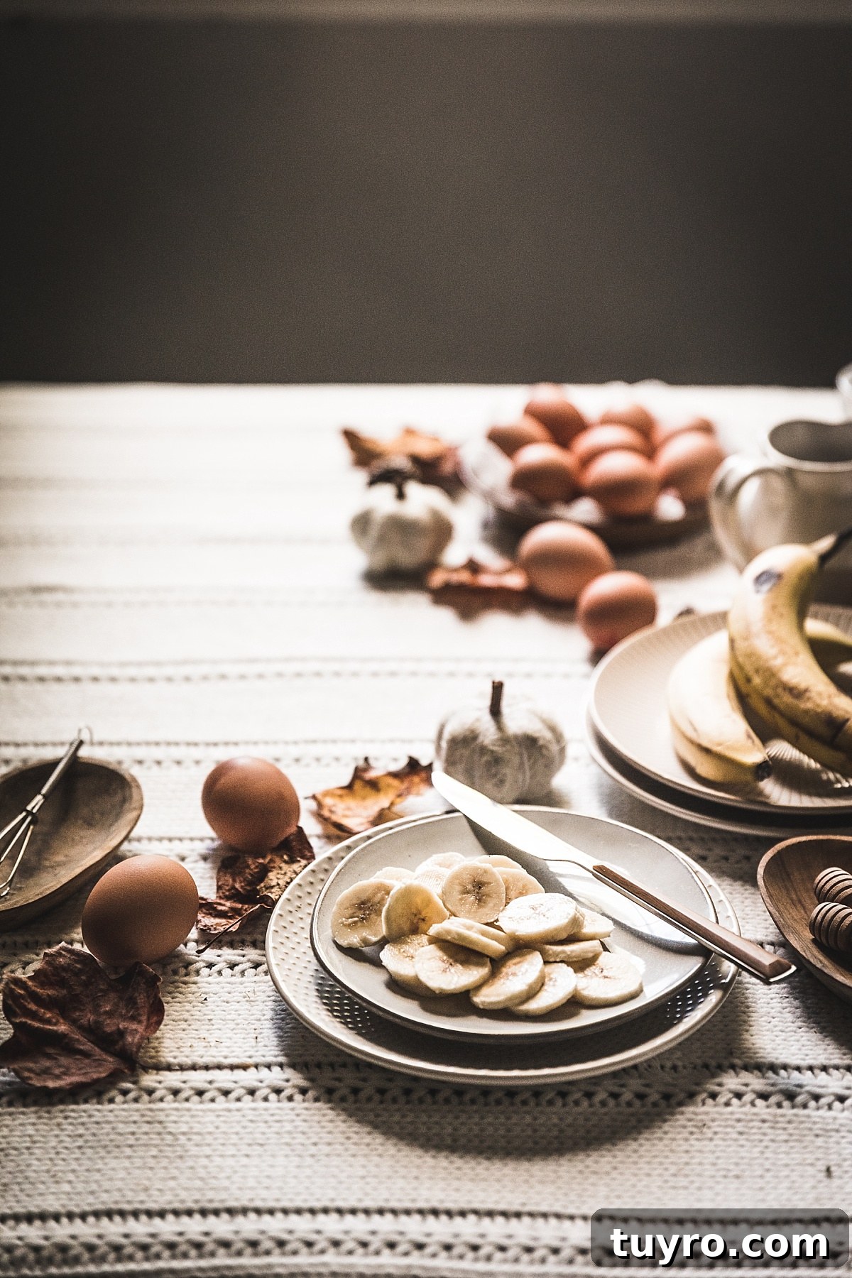2-Ingredient Banana Pancakes: Egg & Banana Recipe 8 high contrast moody autumn image of sliced bananas surrounding by brown leaves, brown eggs, and pancake toppings