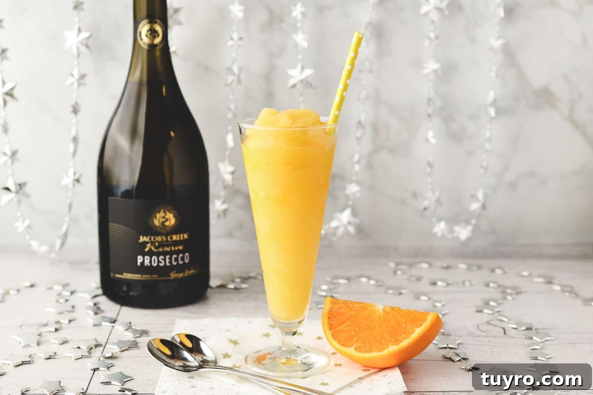 Glass of Mimosa slushy with tall spoons and fresh orange slice on the side.