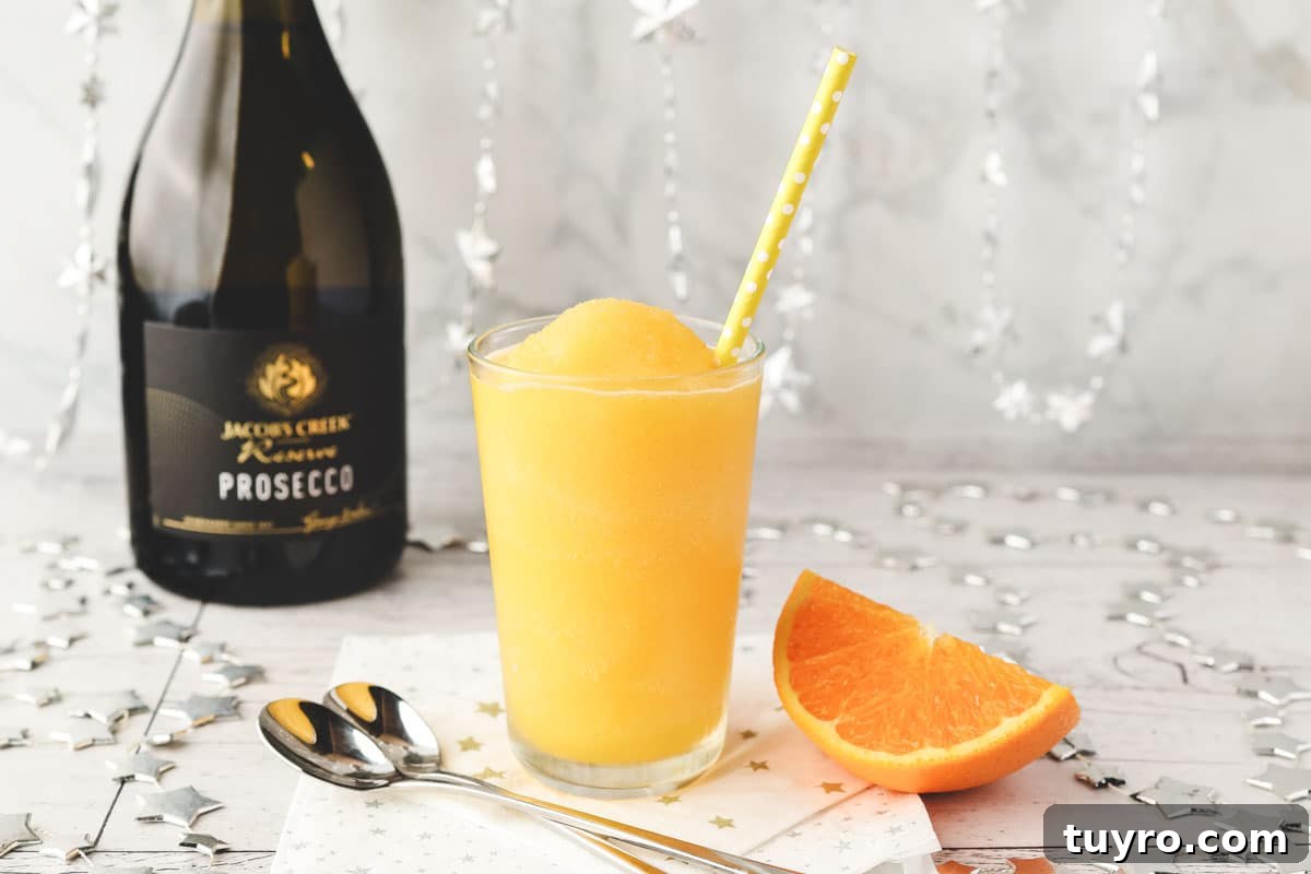 Glass of Mimosa slushy with tall spoons and fresh orange slice on the side.