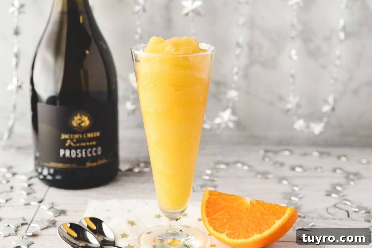 Glass of Mimosa slushy with tall spoons and fresh orange slice on the side.