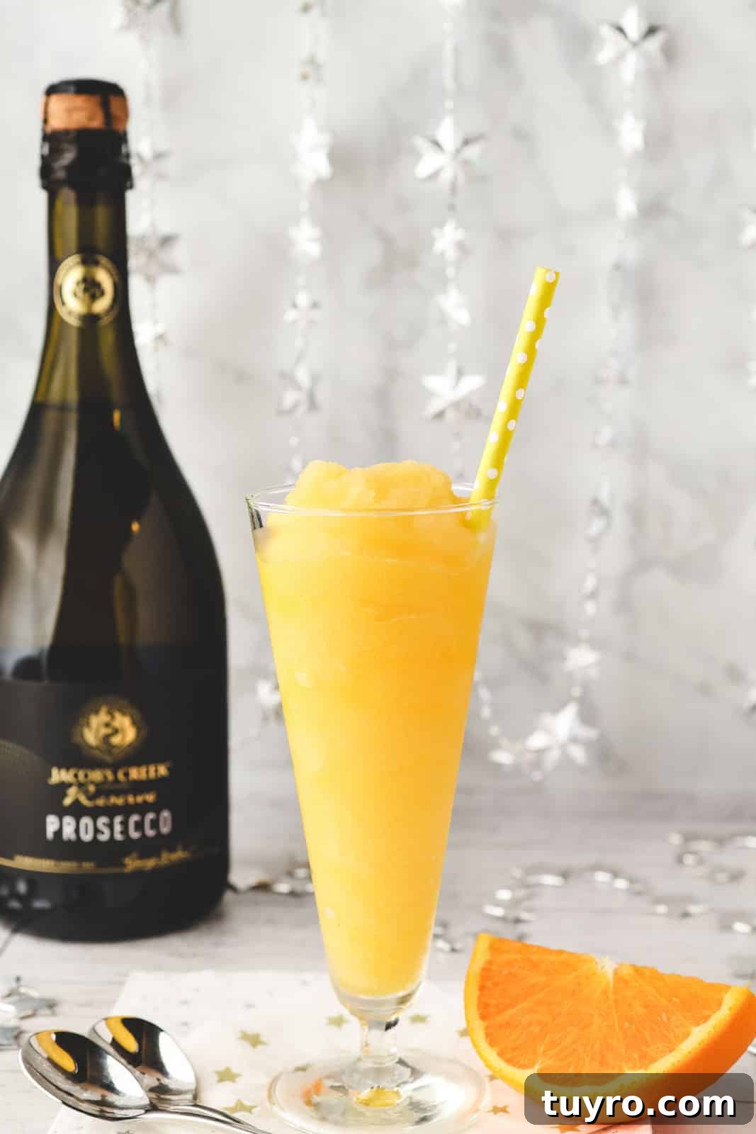 Glass of Mimosa slushy with tall spoons and fresh orange slice on the side.