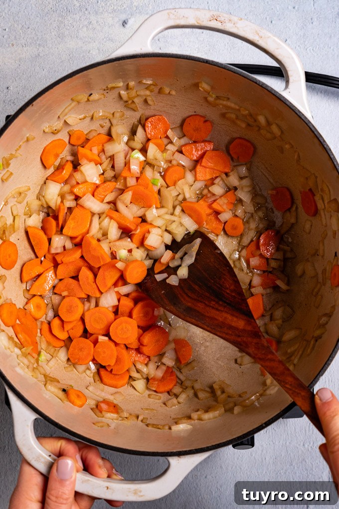 Creamy Broccoli Cheddar Soup Recipe with Gluten-Free Option 4 Overhead shot of carrots and onions being sautéed in a white Dutch oven. A hand is holding one of the handles of the Dutch oven while another hand is holding a wooden spoon and is stirring the ingredients in the pot. #foodphotography