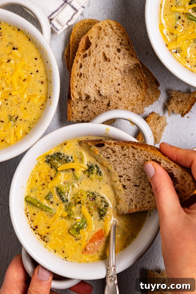 Creamy Broccoli Cheddar Soup Recipe with Gluten-Free Option 9 Overhead photo of healthy broccoli cheddar soup in a white bowl. A hand is holding a piece of bread and dipping it into the soup. There is a bowl of soup in the upper left corner, and another bowl of soup in the upper right corner. More slices of bread are scattered through the scene. #foodphtoography