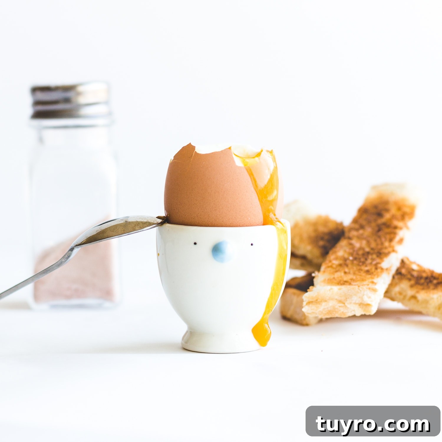 How to Make Creamy Soft-Boiled Eggs Every Time 7 Soft boiled egg in egg cup with top off, yolk running down the side and spoon leaning on the edge. Pile of toast soldiers and salt shaker in the background.