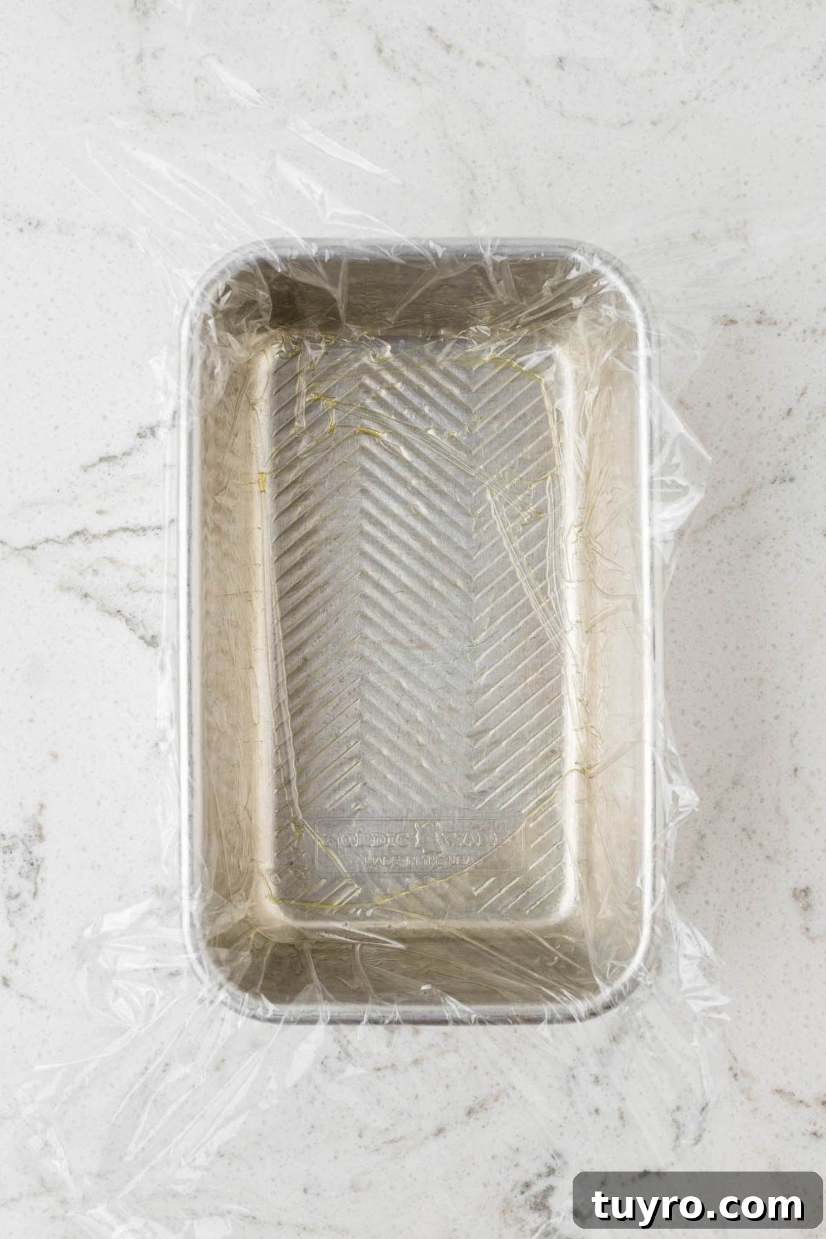 A metal loaf pan lined with plastic wrap sits on a white marbled surface.