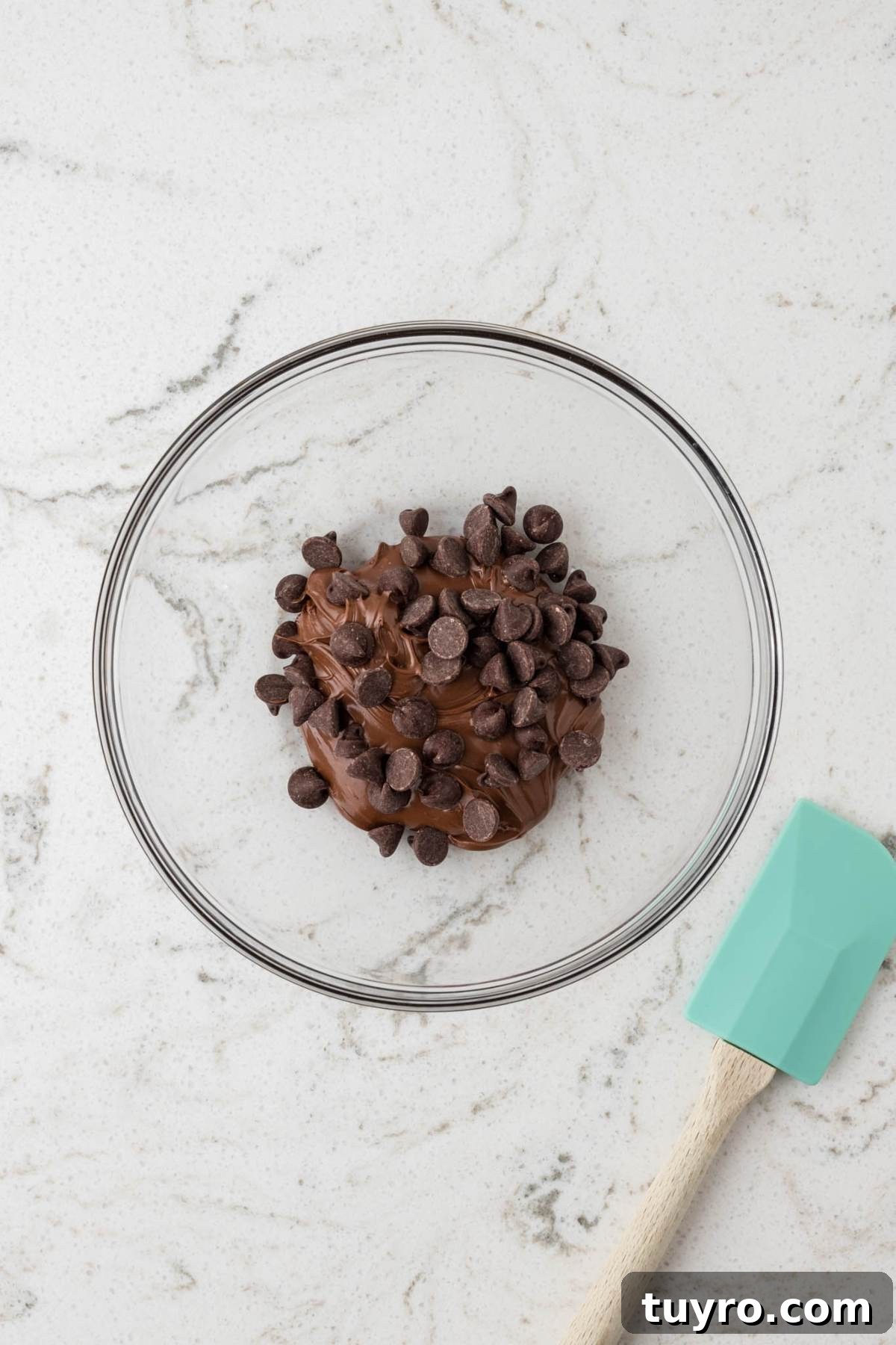 A glass bowl with chocolate chips and spread, next to a blue spatula on a marble countertop.
