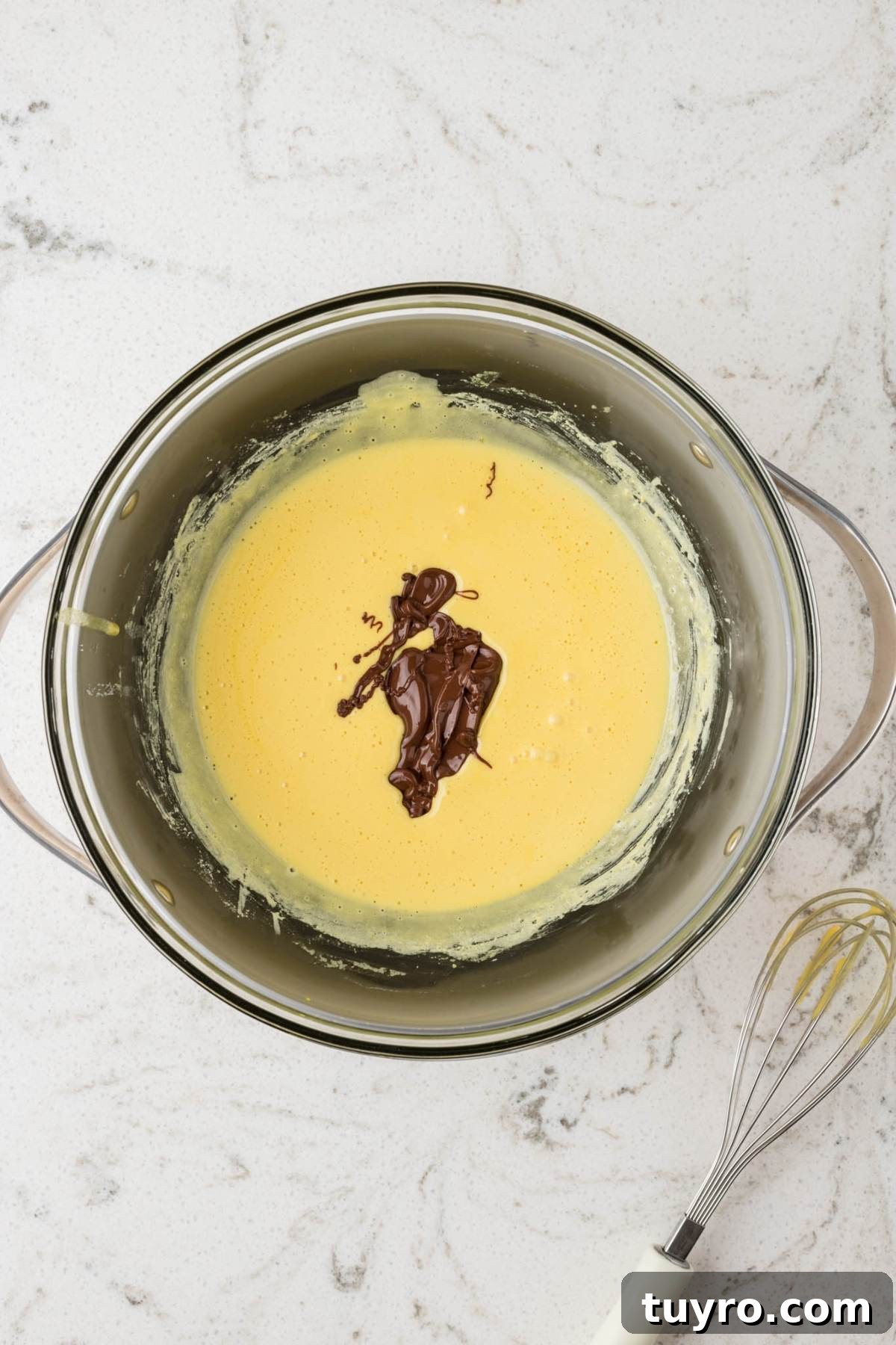 A metal bowl with yellow batter and melted chocolate, next to a whisk on a white countertop.