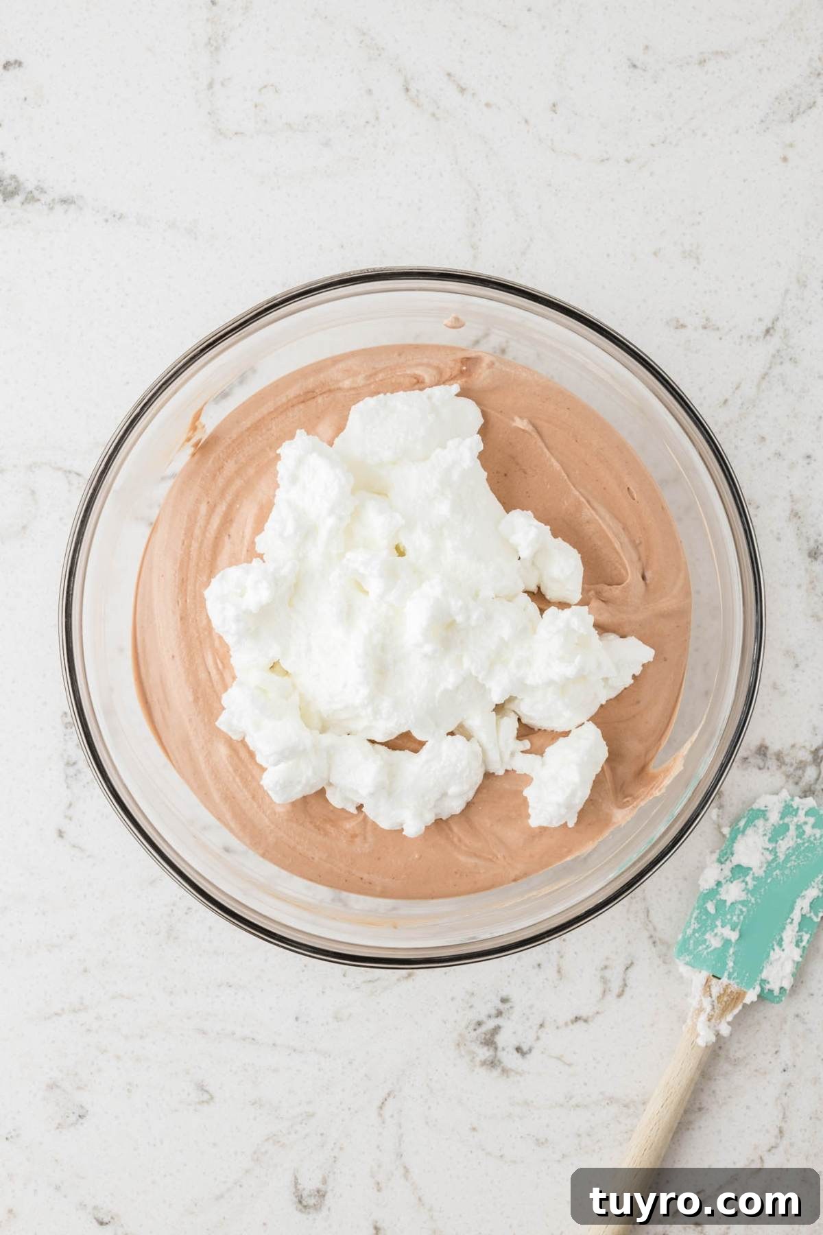 A glass bowl with chocolate mousse and whipped egg whites next to a green spatula on a marble surface.