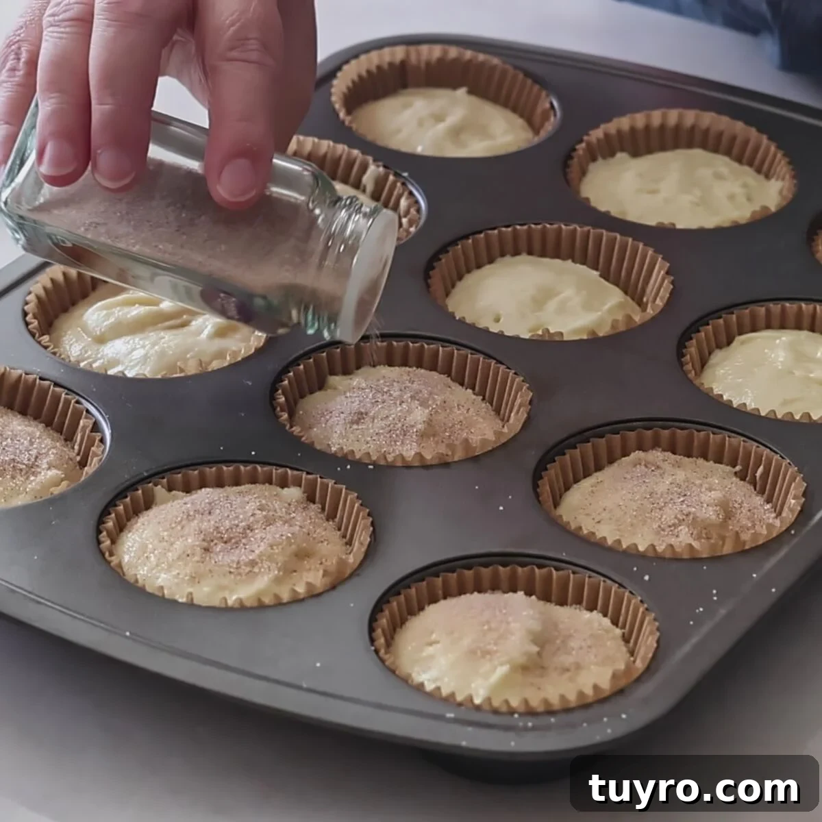 sprinkling cinnamon and sugar on top of cupcake batter in muffin pan.