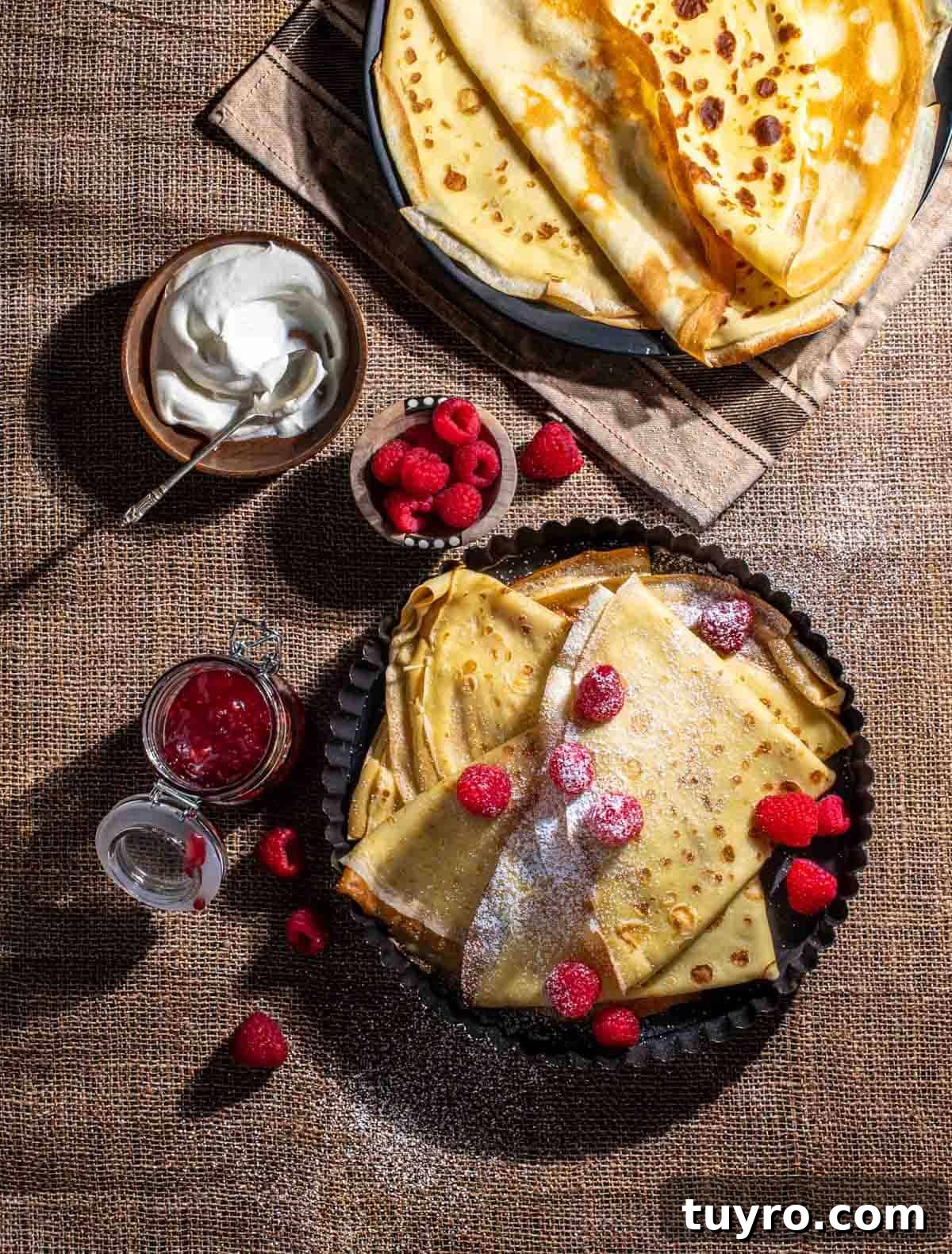 Danish Pandekager: Crispy-Edged Pancakes Recipe 2 Pandekager folded in triangles in a black, round backing dish, served with raspberries, icing sugar, jam and cream.