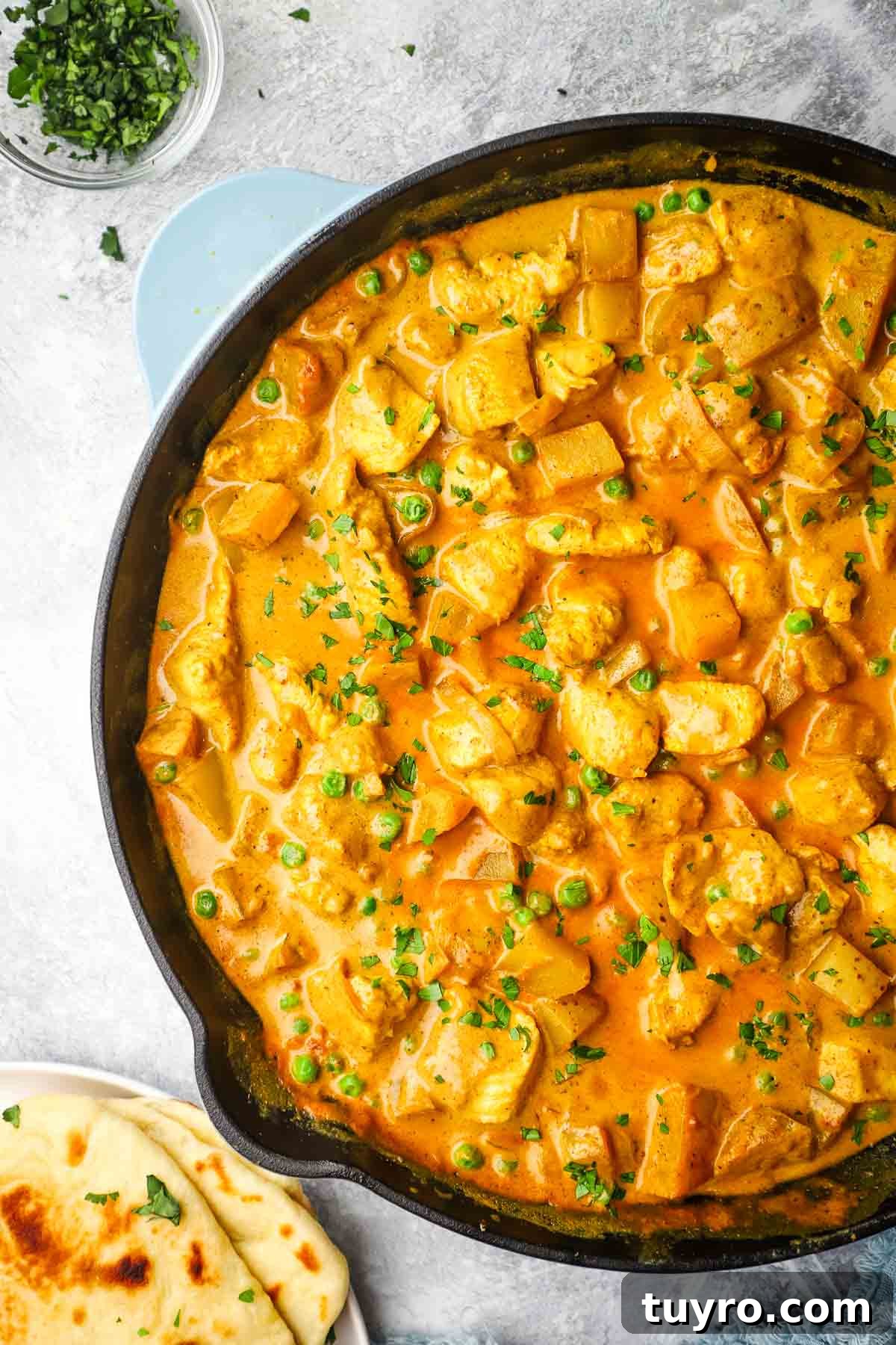 Chicken and Potato Curry Recipe 18 A cast iron pan full of chicken and potato curry garnished with cilantro, with a plate of naan in the background.