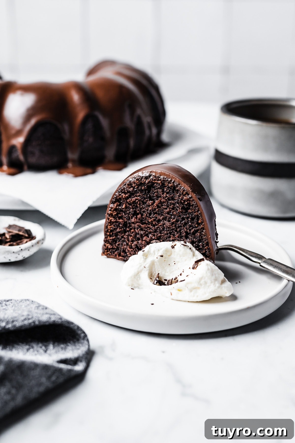 Slice of chocolate olive oil bundt cake with chocolate ganache glaze on a white plate with coffee mug and cake in background
