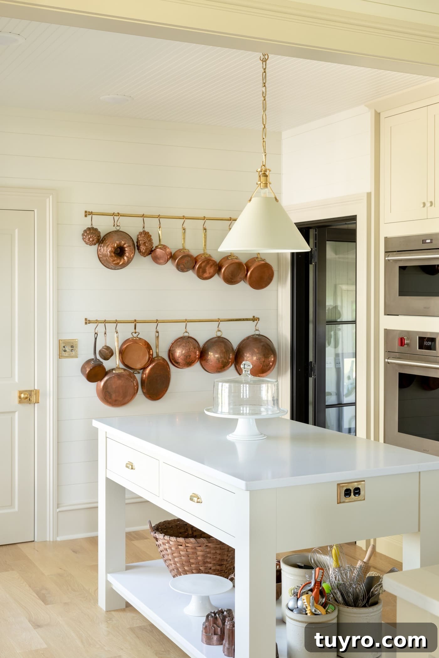 Small island with chunky legs and lower shelf covered in marble with wall oven behind and rack filled with copper cookware