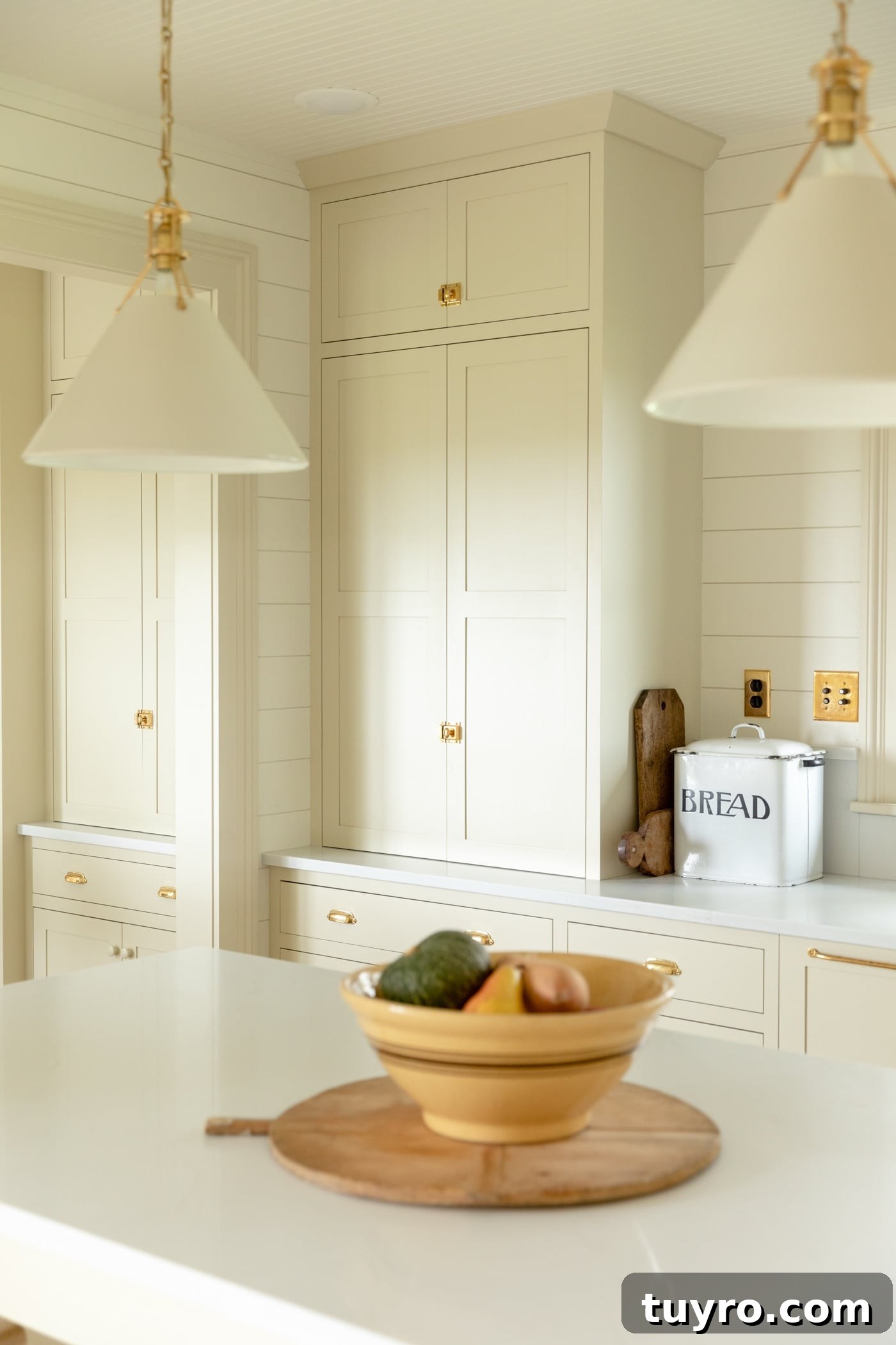 Large pantry cabinet in creamy white farmhouse kitchen with yellow bowl filled with squash sitting on large island with white countertop and pendants hanging above island