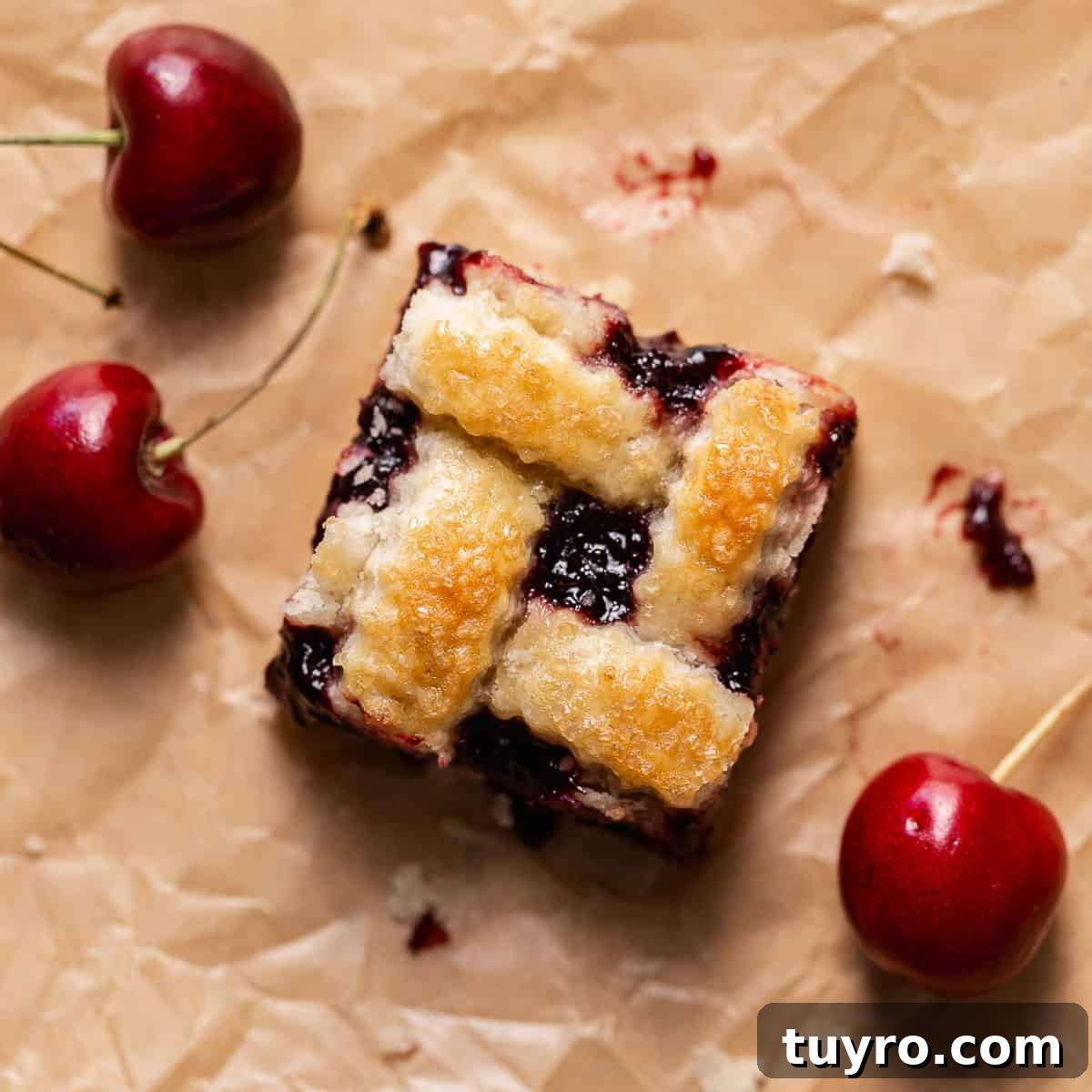 Homemade Cherry Pie Bars with Buttery Crust 6 A cherry pie bar on brown parchment paper surrounded by fresh cherries.