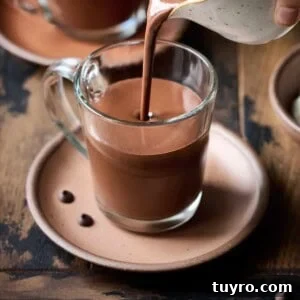 Creamy Vegan Hot Chocolate Recipe 14 Person pouring hot chocolate into a glass mug on a wood table.