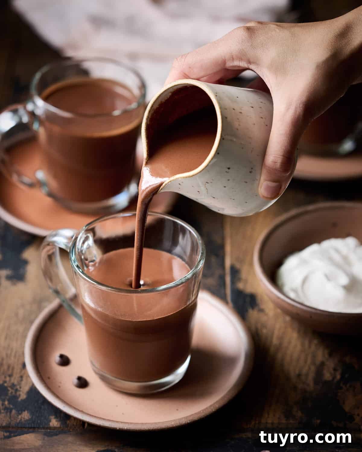 Creamy Vegan Hot Chocolate Recipe 5 Person pouring hot chocolate into glass mug on wood table.