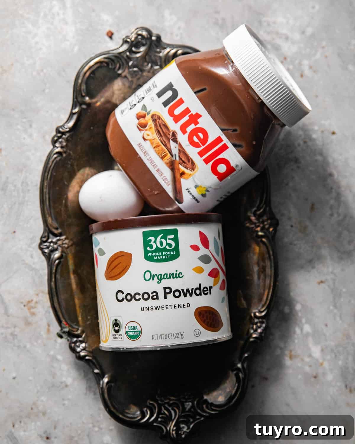 3-Ingredient Mug Cake Recipe Ready in Minutes 3 ingredients to make 3 ingredient mug cake on a grey surface - nutella, cocoa powder, and an egg.