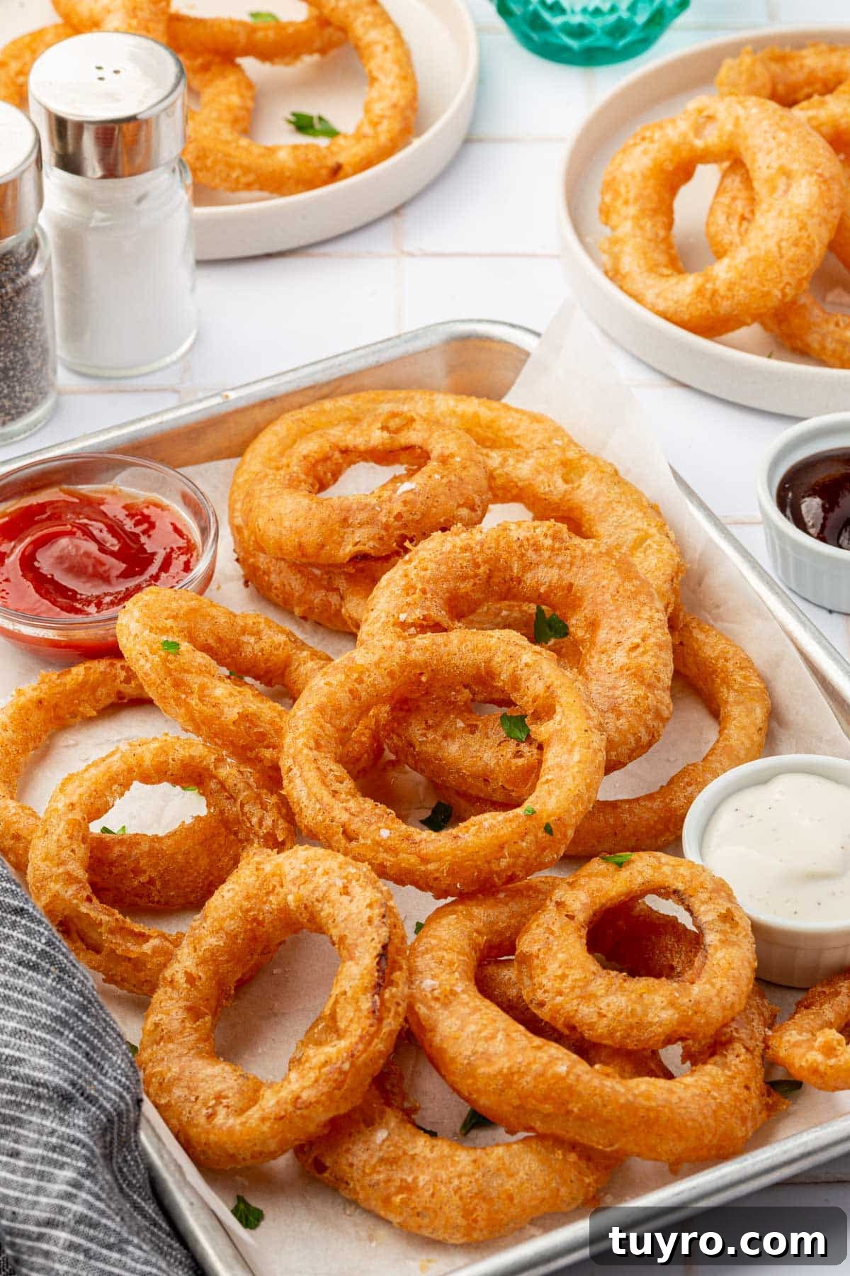 Crispy Gluten-Free Onion Rings Recipe 12 A parchment lined baking sheet of gluten free onion rings with ramekeins of ketchup and ranch with additional plates of onion rings in the background.