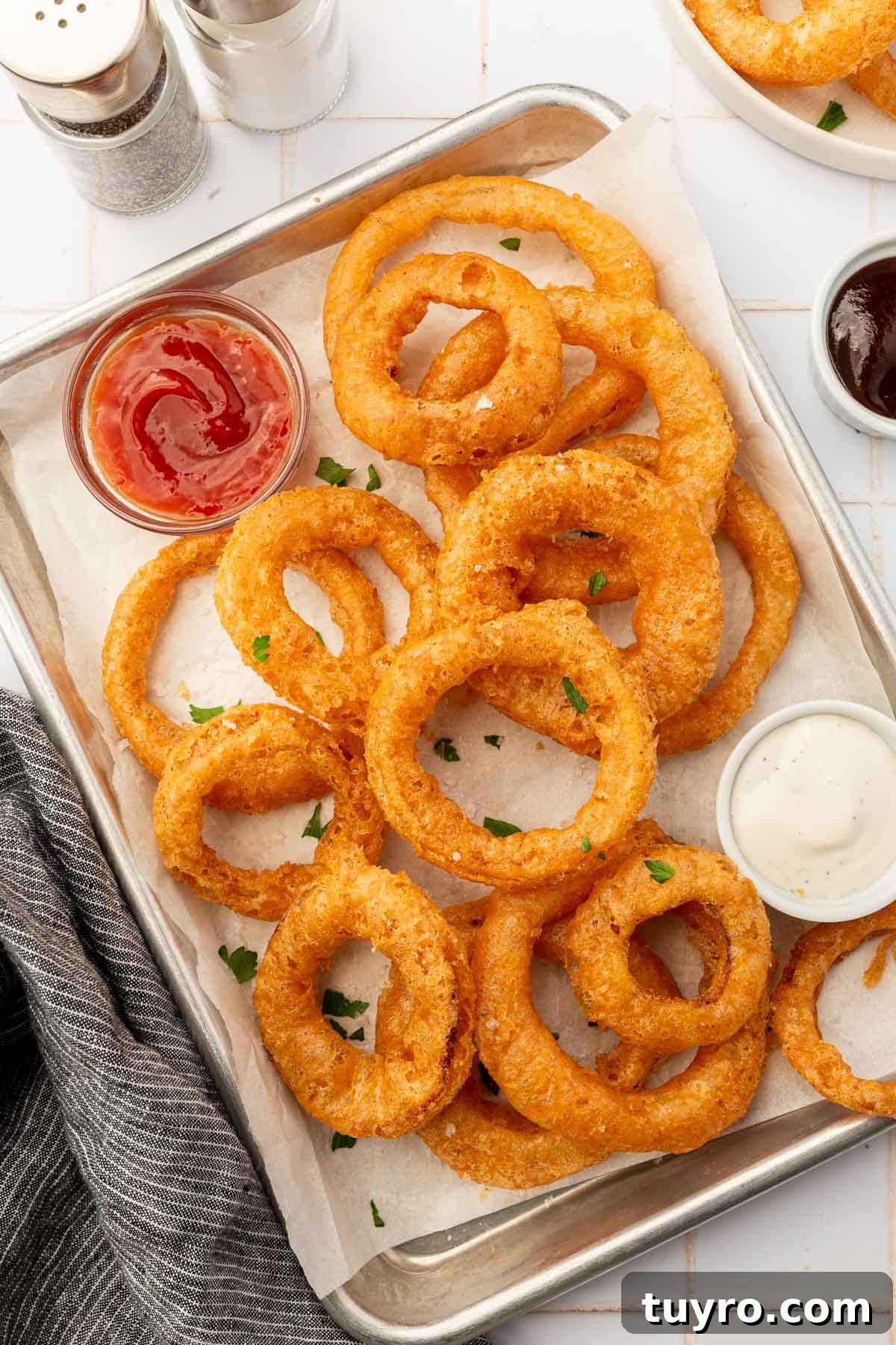 Crispy Gluten-Free Onion Rings Recipe 13 Gluten-free onion rings on a parchment lined baking sheet with a ramekin of ranch and a ramekin of ketchup.