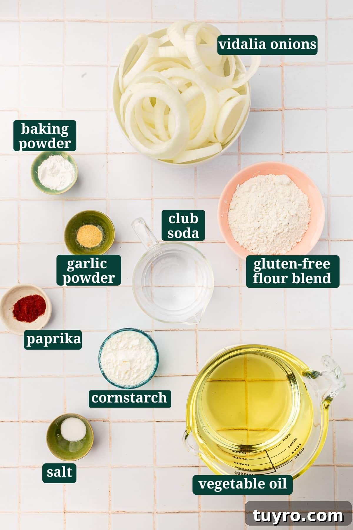 Crispy Gluten-Free Onion Rings Recipe 3 An overhead view of ingredients in bowls to make gluten free onion rings, including vidalia onions, baking powder, garlic powder, paprika, club soda, cornstarch, gluten-free flour blend, vegetable oil and salt with text overlays over each ingredient.