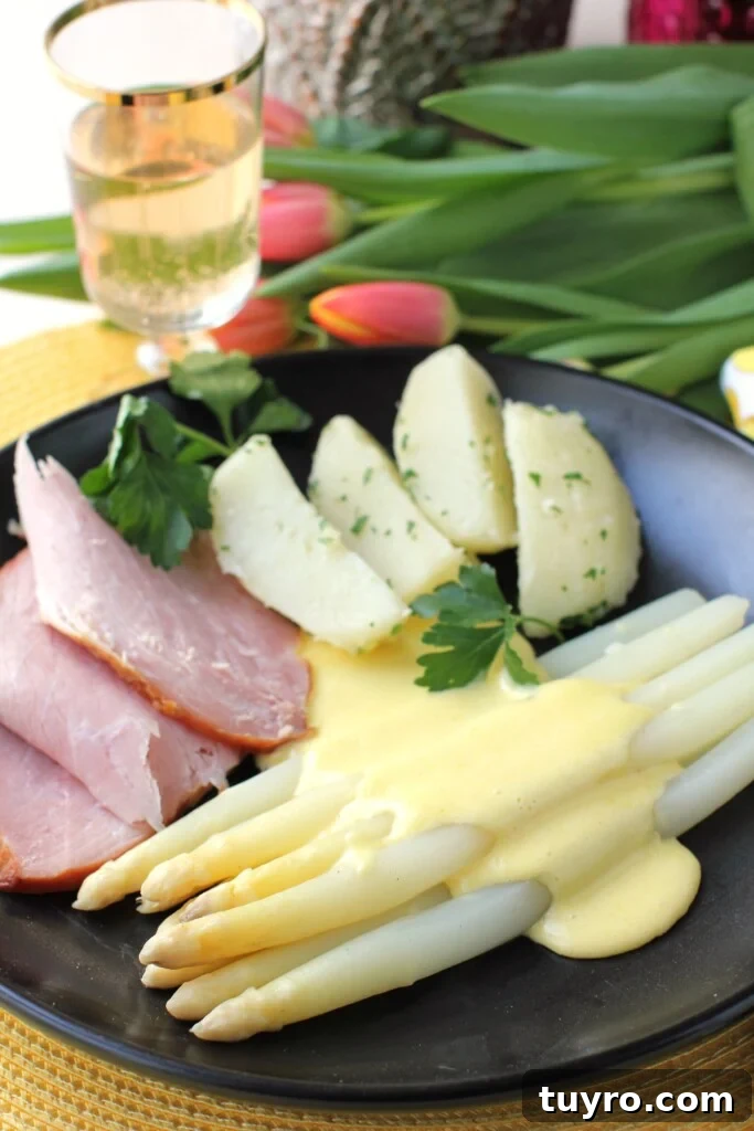Classic Hollandaise Sauce Recipe for White Asparagus 2 A black plate with ham, boiled potatoes, and white asparagus topped with Easy Hollandaise Sauce; glass of wine and pink tulips in the background.
