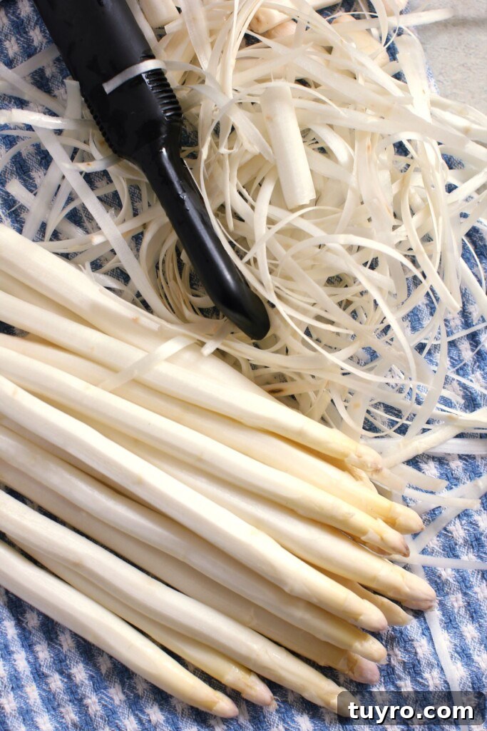 Classic Hollandaise Sauce Recipe for White Asparagus 4 Peeled white asparagus stalks on a blue towel, with a pile of peelings beside and a black vegetable peeler.