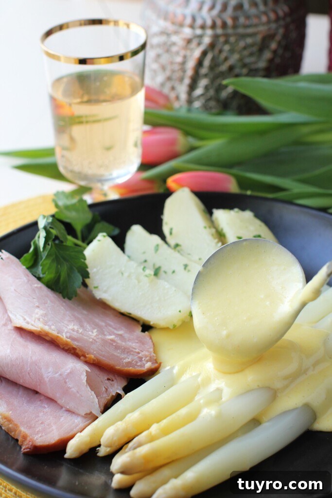 Classic Hollandaise Sauce Recipe for White Asparagus 6 Ladling Hollandaise sauce over cooked white asparagus on a black plate with sliced ham and boiled potatoes.