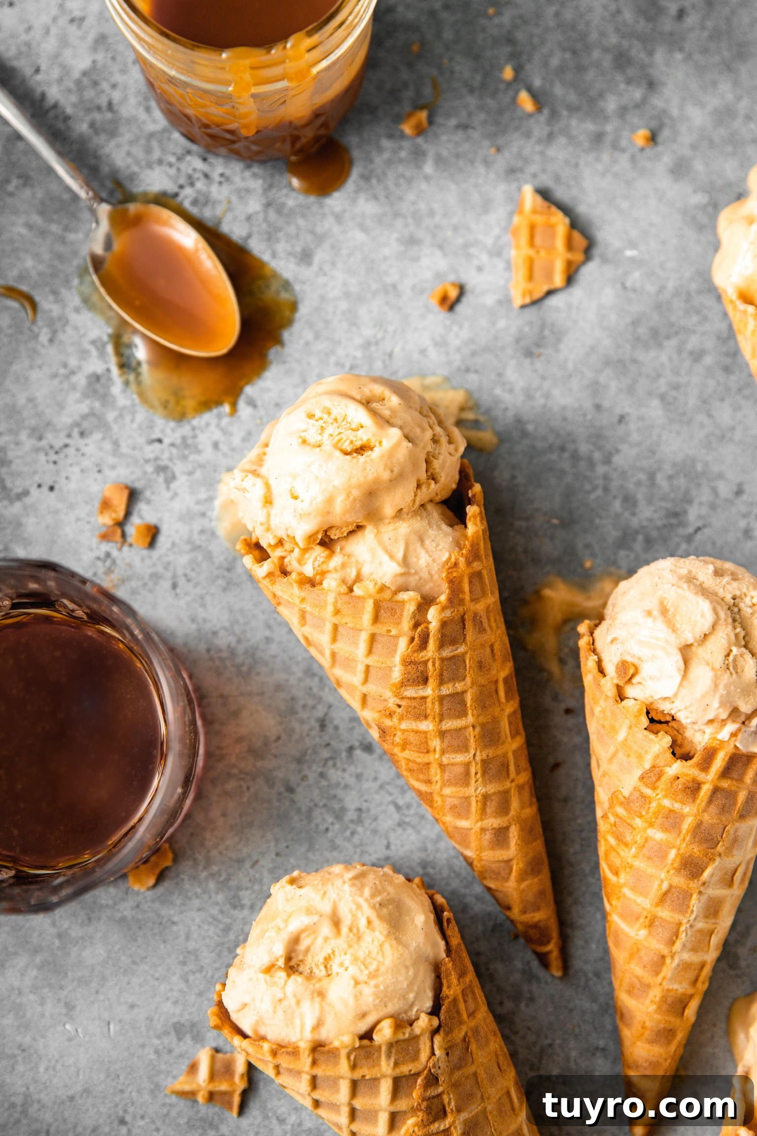 No-Churn Bourbon Salted Caramel Ice Cream Recipe 7 Close up of a double scoop of bourbon salted caramel ice cream inside of an ice cream cone with a glass of bourbon and mason jar of bourbon caramel nearby.