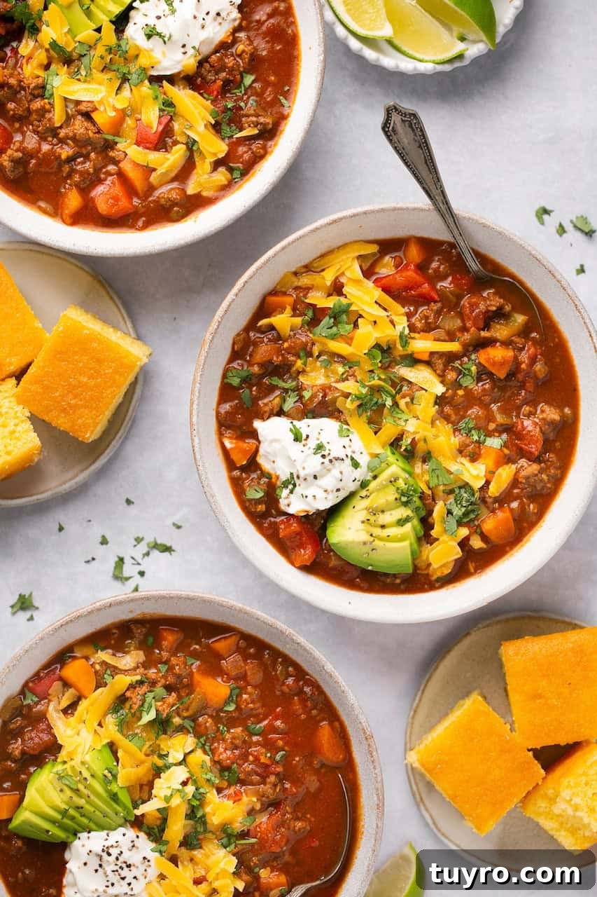 Hearty No-Bean Chili Recipe for Rich, Meaty Flavor 2 no bean chili in 3 bowls topped with cheese, sour cream, and avocado with cornbread on the side