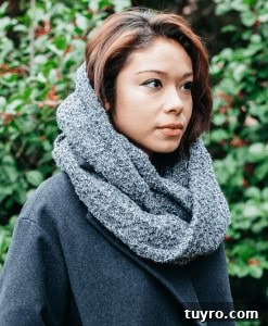 Image shows a young woman with dark hair standing in front of greenery wearing a large cowl neck scarf.