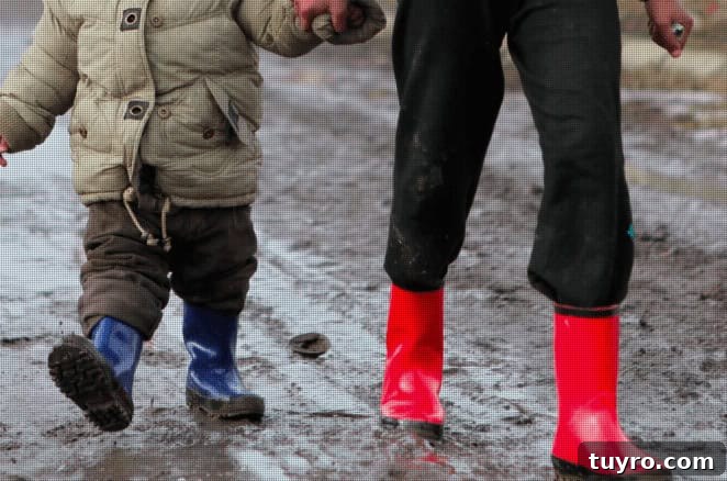 Image shows the bottom half of two young children wearing brightly colored rainboots, walking through mud puddles