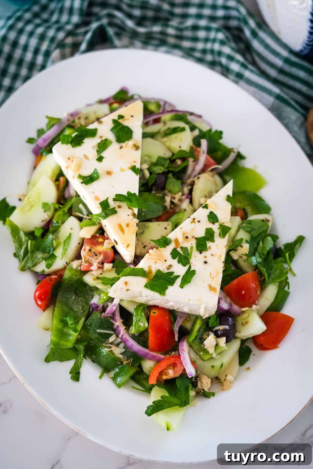 Authentic Greek Horiatiki Salad Recipe — Classic Village Salad 2 A plate of Greek salad with sliced cucumbers, tomatoes, red onions, and green peppers, topped with two triangular pieces of feta cheese and garnished with parsley.