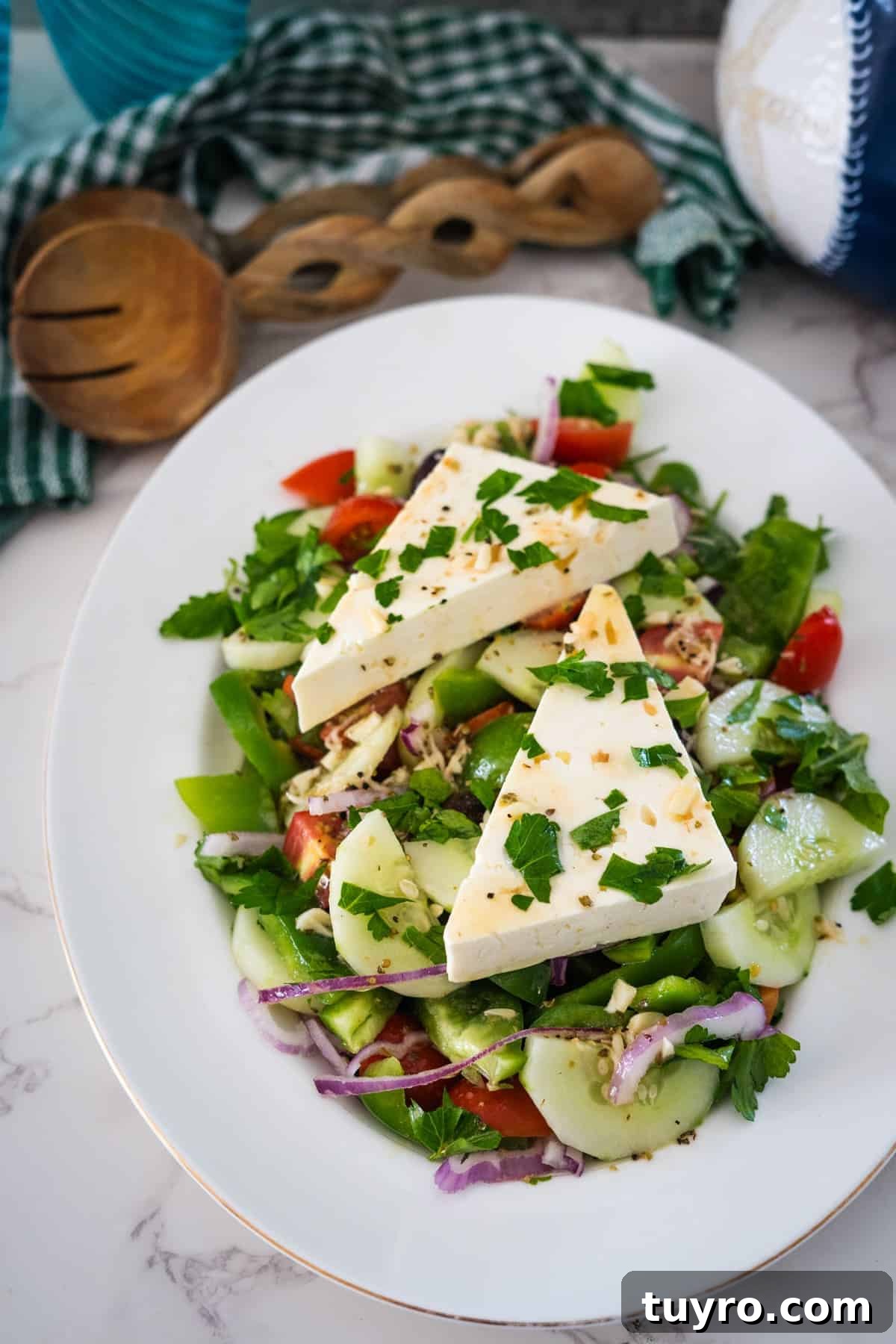 Authentic Greek Horiatiki Salad Recipe — Classic Village Salad 11 A Greek village salad with cucumbers, tomatoes, red onions, green peppers, and parsley, topped with two large slices of cheese. It's served on a white plate with wooden utensils and a green checkered cloth in the background.