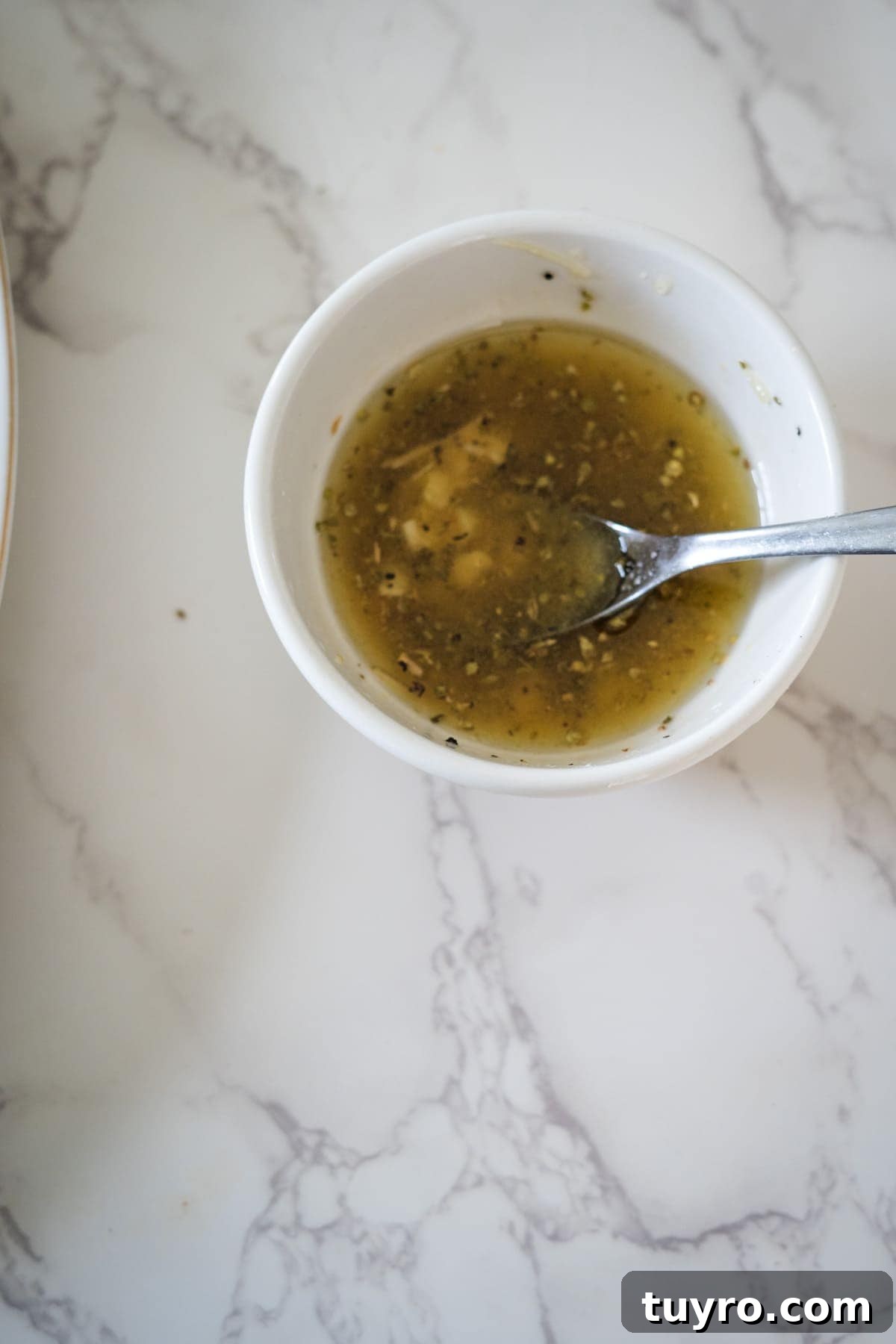 Authentic Greek Horiatiki Salad Recipe — Classic Village Salad 4 A round white bowl containing a light brown liquid with tiny chopped ingredients, reminiscent of a Greek village salad, and a spoon inside, resting on a white marbled surface.