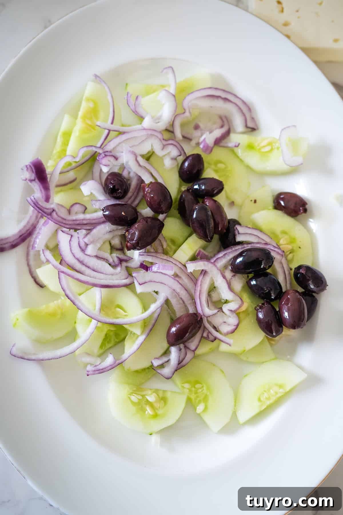 Authentic Greek Horiatiki Salad Recipe — Classic Village Salad 5 A white plate with sliced cucumbers, red onion, and black olives arranged on it, reminiscent of a classic Greek village salad.