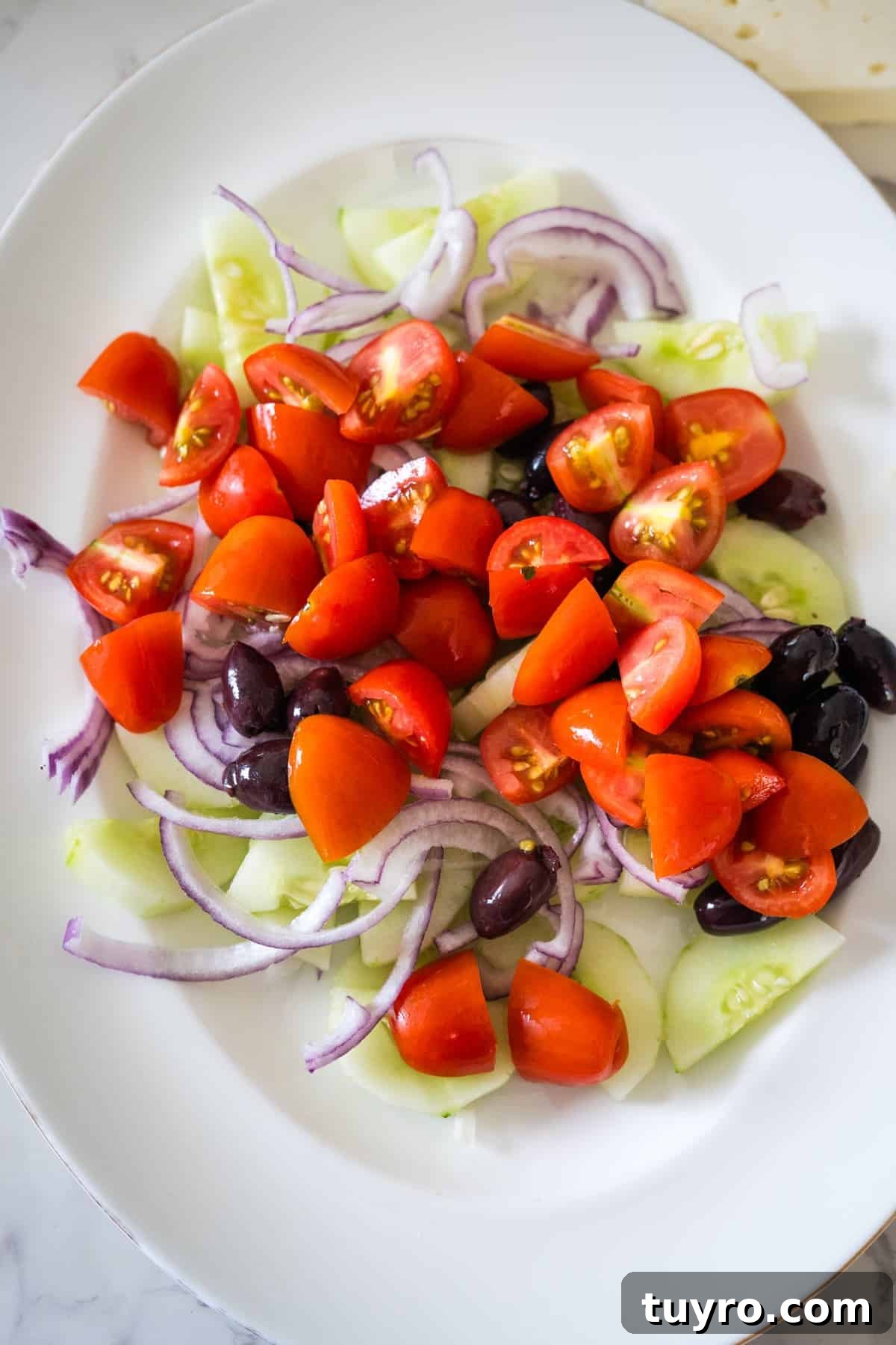 Authentic Greek Horiatiki Salad Recipe — Classic Village Salad 6 A white plate holds a Greek village salad featuring fresh chopped cherry tomatoes, sliced cucumbers, red onion rings, and black olives.